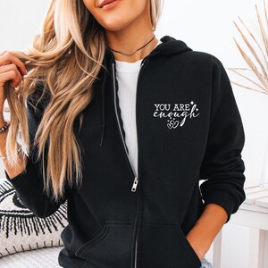 May include: Black zip-up hoodie with the text "YOU ARE enough" in white script. The hoodie has a drawstring hood, a full-length zipper, and two front pockets. The design includes two hearts and a star.