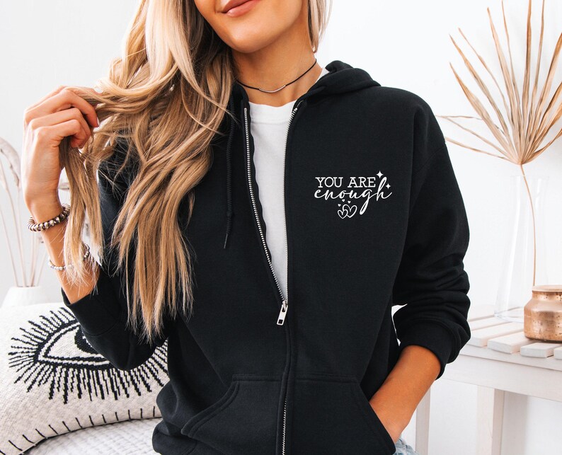 May include: Black zip-up hoodie with the text "YOU ARE enough" in white script. The hoodie has a drawstring hood, a full-length zipper, and two front pockets. The design includes two hearts and a star.