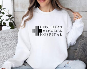 Puede incluir: Una sudadera blanca de manga larga con cuello redondo y un gráfico negro en el pecho. El gráfico muestra un símbolo de cruz estilizado junto al texto negro que dice GREY + SLOAN MEMORIAL HOSPITAL en una fuente sans-serif limpia.