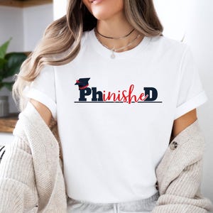 May include: A white short-sleeved t-shirt featuring the text PhinisheD in black and red lettering. The letter P is topped with a black graduation cap with a red tassel. The design is centred on the chest of the shirt, which is worn with a cardigan.