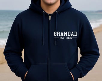 Puede incluir: Sudadera con capucha azul marino con el texto blanco "GRANDAD EST 2026". La sudadera con cremallera tiene capucha, cremallera completa y dos bolsillos delanteros. El fondo muestra una playa y el mar.