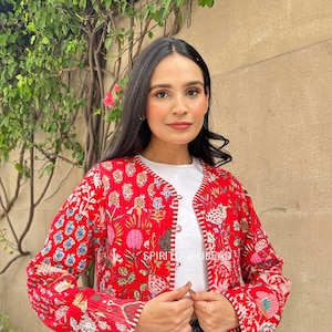 Może przedstawiać: Czerwona pikowana kurtka z kwiatowym nadrukiem i białym wykończeniem. Kurtka zapinana jest na guziki i ma długie rękawy. Modelka nosi kurtkę na białą koszulę i niebieskie dżinsy.