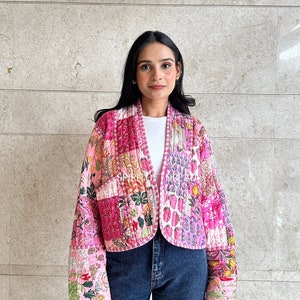 May include: A pink quilted jacket with a floral print. The jacket has a cropped fit and long sleeves. The model is wearing the jacket with blue jeans.