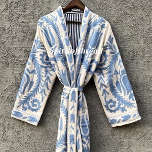 May include: A long, cream-colored robe with intricate blue floral embroidery. The robe has long sleeves and a matching belt. The interior lining is striped in black and white. The text "Spiritofthread" is visible on the robe.