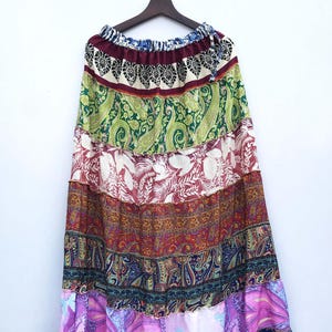 以下が含まれることがあります： さまざまな柄のカラフルな生地を使った、ロング丈のティアードスカート。ペイズリー柄、花柄、抽象柄が緑、赤、紫、青の色合いでデザインされています。白い背景に、木製のハンガーに吊るされています。