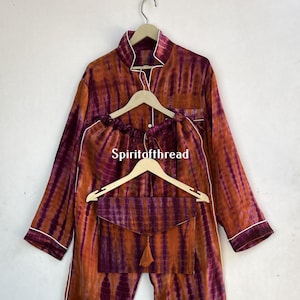 Puede incluir: Un conjunto de pijama de teñido anudado con una parte superior de manga larga y pantalones. El conjunto está en tonos de naranja, morado y blanco. La parte superior tiene un cuello y un cierre de botón. Los pantalones tienen una cintura elástica. El conjunto está colgado en una percha de madera. El texto "Spiritofthread" es visible en la percha.