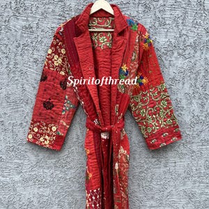 May include: A long-sleeved red quilted robe featuring a patchwork design with various floral and geometric patterns in contrasting colors. The garment includes a notched collar, a tie-waist belt, and the text Spiritofthread printed on the front.