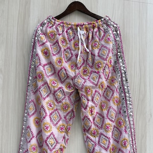 May include: Light pink trousers featuring a repeating pattern of yellow and magenta embroidered diamonds and circular motifs. The pants include a white drawstring waist and vertical side panels decorated with small reflective silver mirrors.