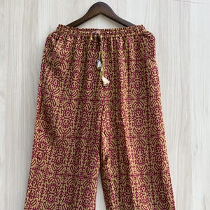 May include: Wide-leg pants featuring a repeating maroon and tan geometric pattern. The waistband is elasticized with a drawstring closure. The hem is finished with a horizontal border displaying a stylized blue and white fish motif on a tan background.