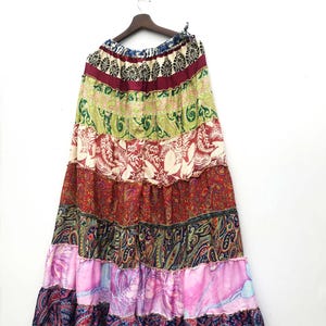 May include: A long, tiered skirt with a patchwork design. The skirt features various patterns and colors, including paisley, floral, and abstract prints. The skirt is hanging on a wooden hanger against a white background.
