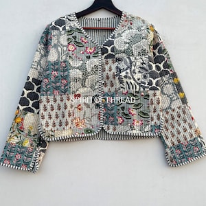 Może przedstawiać: Kurtka pikowana w stylu patchwork z dekoltem w kształcie litery V i zapięciem na guziki. Kurtka jest wykonana z bawełny i zdobiona różnymi wzorami kwiatowymi i geometrycznymi w odcieniach niebieskiego, zielonego, białego i czarnego. Na przodzie kurtki widoczny jest napis "SPIRIT OF THREAD".