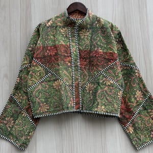 May include: A quilted jacket featuring a green and red floral pattern with intricate paisley details. The garment has a high collar, long sleeves, and a front opening trimmed with a black and white striped border that defines the seams.