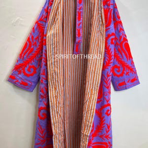 May include: A long, open-front robe with a vibrant design. The outer layer is a solid purple with bold red embroidered floral patterns. The interior features orange and white vertical stripes. The robe is hanging on a wooden hanger.
