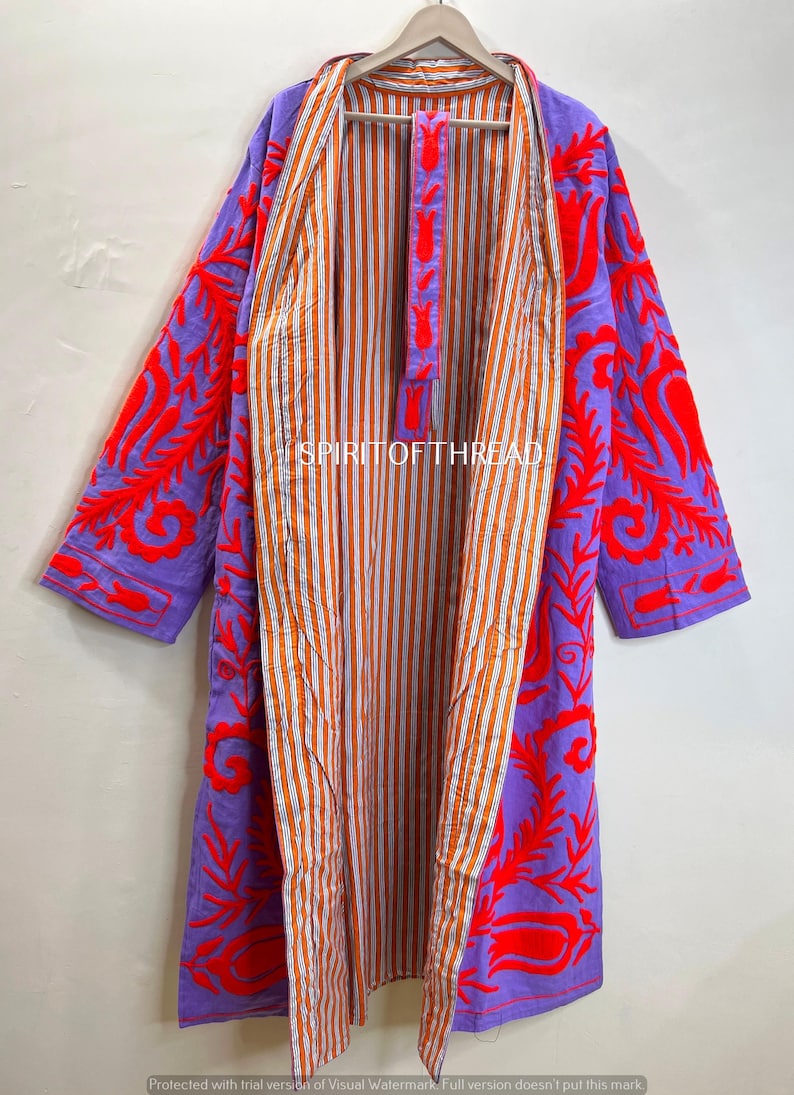 May include: A long, open-front robe with a vibrant design. The outer layer is a solid purple with bold red embroidered floral patterns. The interior features orange and white vertical stripes. The robe is hanging on a wooden hanger.