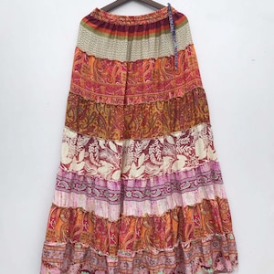 May include: A long, tiered skirt with a vibrant, patchwork design. The skirt features a variety of patterns, including paisley and floral prints, in shades of red, orange, pink, and white. It hangs from a wooden hanger, showcasing its flowing, bohemian style.