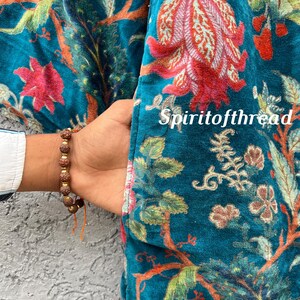 Pode incluir: Casaco de veludo azul-petr&oacute;leo com um padr&atilde;o floral em tons de vermelho, laranja e verde. O casaco tem um bolso e est&aacute; parcialmente aberto, revelando uma manga de camisa branca. O texto "Spiritofthread" &eacute; vis&iacute;vel.