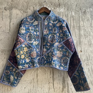 May include: A quilted jacket featuring a blue floral pattern with contrasting dark maroon and green patchwork sections. The garment has a high collar, long sleeves, and white and blue striped piping along the edges and seams, hanging on a hanger.