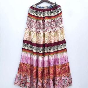 以下が含まれることがあります： さまざまなカラフルな模様とデザインが施された、長いティアードスカート。ピンク、赤、オレンジ、クリームの色合いで、花柄、ペイズリー柄、幾何学模様がミックスされています。白い背景に木製のハンガーから吊り下げられています。