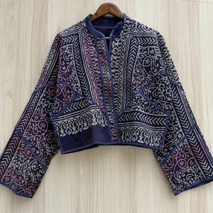 May include: A cropped jacket featuring a deep blue and purple base with intricate white geometric and floral patterns. The garment has long, wide sleeves, a collarless open front, and visible quilted stitching throughout the fabric construction.