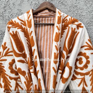 Puede incluir: Una bata blanca con intrincados bordados florales en color naranja. La bata presenta un interior a rayas marr&oacute;n y blanco y est&aacute; colgada de una percha de madera. El texto "Spiritofthread" es visible en la bata.