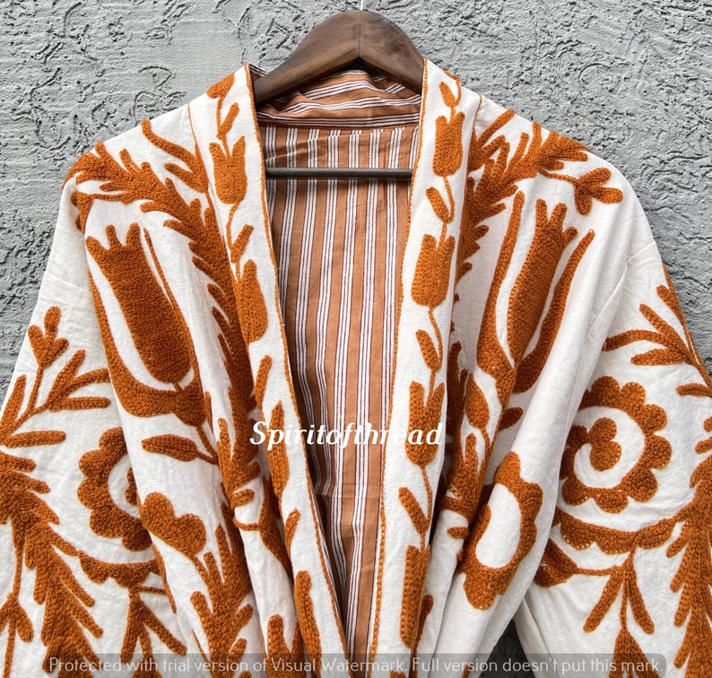 Puede incluir: Una bata blanca con intrincados bordados florales en color naranja. La bata presenta un interior a rayas marr&oacute;n y blanco y est&aacute; colgada de una percha de madera. El texto "Spiritofthread" es visible en la bata.