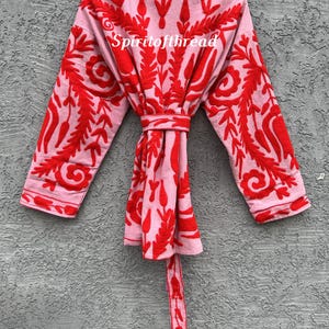 May include: A light pink kimono-style robe with intricate red embroidered floral patterns. The robe has long sleeves and a matching belt. The text "Spiritofthread" is visible on the front of the robe.