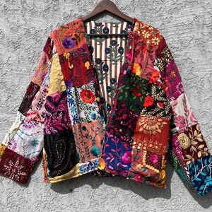 Op de afbeelding: Een patchwork jas met een levendige mix van kleuren en patronen. De jas heeft geborduurde bloemenontwerpen en een gestreepte binnenvoering. De jas wordt getoond op een houten hanger tegen een grijze muur.