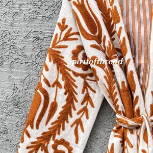 Puede incluir: Bata blanca con intrincado bordado floral de color &oacute;xido. La bata presenta un interior a rayas y un cintur&oacute;n a juego. El texto "Spiritofthread" es visible en la bata. La bata est&aacute; colgada de una percha de madera.