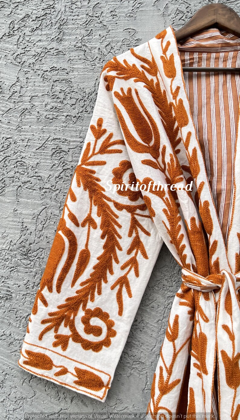 Puede incluir: Bata blanca con intrincado bordado floral de color &oacute;xido. La bata presenta un interior a rayas y un cintur&oacute;n a juego. El texto "Spiritofthread" es visible en la bata. La bata est&aacute; colgada de una percha de madera.