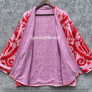 May include: A pink open-front jacket with red floral embroidery on the sleeves and collar. The inside lining features a pink and white striped pattern. The text "Spiritofthread" is visible on the jacket.