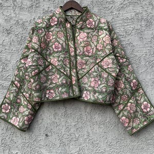 May include: A quilted jacket featuring a green base with a pink floral pattern, a high collar, and long sleeves. The garment is constructed with visible seam lines across the front and sleeves, finished with green piping along the edges.