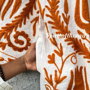 Puede incluir: Primer plano de tela blanca con intrincado bordado naranja. El dise&ntilde;o presenta patrones florales y de hojas, con el texto "Spiritofthread" visible. La tela es sostenida por una mano, mostrando la textura y el detalle del bordado.