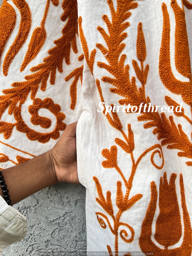 Puede incluir: Primer plano de tela blanca con intrincado bordado naranja. El dise&ntilde;o presenta patrones florales y de hojas, con el texto "Spiritofthread" visible. La tela es sostenida por una mano, mostrando la textura y el detalle del bordado.