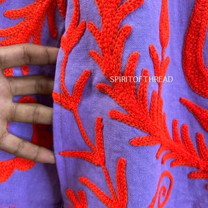May include: Close-up of a lavender fabric with vibrant orange embroidered floral patterns. The design features detailed, stylized leaves and branches. The text "SPIRITOF THREAD" is visible on the fabric.