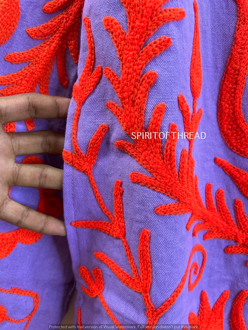 May include: Close-up of a lavender fabric with vibrant orange embroidered floral patterns. The design features detailed, stylized leaves and branches. The text "SPIRITOF THREAD" is visible on the fabric.