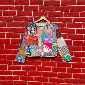Peut inclure: Veste patchwork colorée avec divers motifs floraux et géométriques. La veste, sans col, est présentée sur un cintre en bois devant un mur de briques rouges. Le texte "SPIRIT OF THREAD" est visible sur la veste.