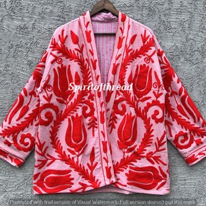 May include: A pink kimono-style jacket with a red floral pattern. The jacket has long sleeves and a relaxed fit. The text "Spiritofthread" is visible on the jacket. The jacket is hanging on a wooden hanger.