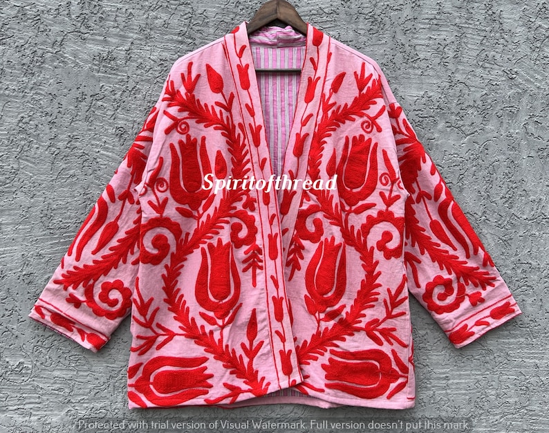 May include: A pink kimono-style jacket with a red floral pattern. The jacket has long sleeves and a relaxed fit. The text "Spiritofthread" is visible on the jacket. The jacket is hanging on a wooden hanger.