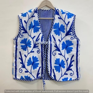 Könnte beinhalten: Eine weiße Weste mit blauer Blumenstickerei und blau-weiß gestreiftem Innenfutter. Die Weste hat einen Kordelverschluss und wird auf einem Holzbügel präsentiert. Das florale Design umfasst blaue Blumen und Stiele.