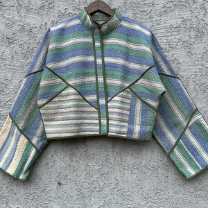 May include: A cropped jacket with a stand-up collar and a zipper closure. The jacket features a patchwork design with horizontal stripes in shades of blue, green, and cream. The seams are outlined in a darker green, and the jacket is hanging on a wooden hanger.