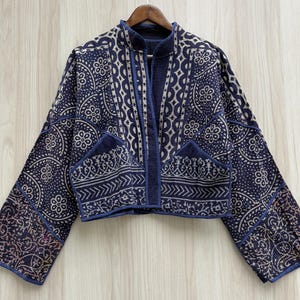 Puede incluir: Una chaqueta corta en azul marino profundo con un patrón de estampado floral y geométrico en color crema. La prenda presenta mangas largas, cuello mao y dos bolsillos delanteros, todo ello rematado con costuras visibles en la tela.