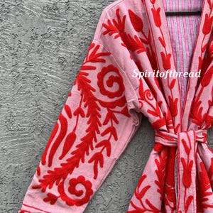 May include: A light pink robe with red floral embroidery. The robe features long sleeves and a tie belt. The interior lining is striped pink and white. The text "Spiritofthread" is visible on the robe.