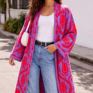 May include: A long, open-front kimono-style jacket in a vibrant purple and red floral pattern. The jacket is worn over a white top and blue jeans. A small white purse hangs from the shoulder.