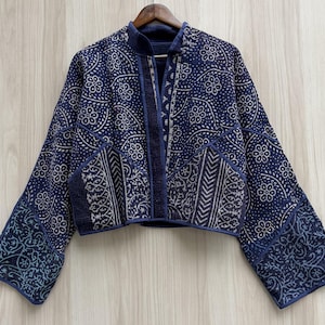 Puede incluir: Una chaqueta corta en azul índigo profundo con un estampado de flores y cachemira en blanco. La prenda tiene mangas largas y anchas, una abertura frontal recta y un cuello tipo mandarín, todo exhibido en una percha de madera.