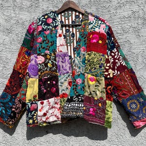 Könnte beinhalten: Eine Patchwork-Jacke mit einer Vielzahl von bunten Blumen- und geometrischen Mustern. Die Jacke hat eine Mischung aus Stoffen und Texturen, einschließlich Stickereien und Samt, und hängt an einem Holzbügel.