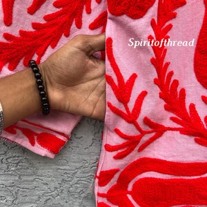 May include: Close-up of a pink fabric with vibrant red embroidered floral patterns. The design features intricate, raised stitching in a bold, eye-catching style. The text "Spiritofthread" is visible on the fabric.