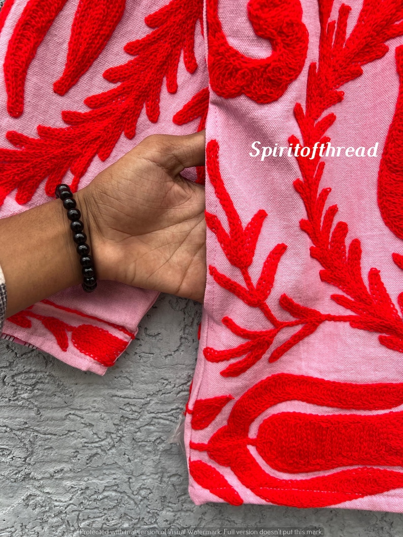 May include: Close-up of a pink fabric with vibrant red embroidered floral patterns. The design features intricate, raised stitching in a bold, eye-catching style. The text "Spiritofthread" is visible on the fabric.
