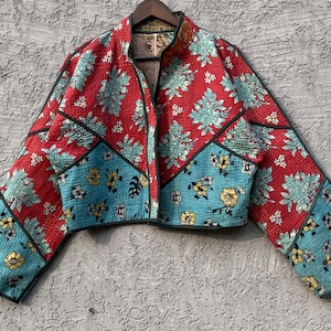 Può includere: Giacca corta con un vivace motivo floreale. La giacca presenta un design patchwork con stampe floreali rosse, turchesi e gialle. I bordi sono delineati da finiture nere. La giacca è esposta su una gruccia di legno.