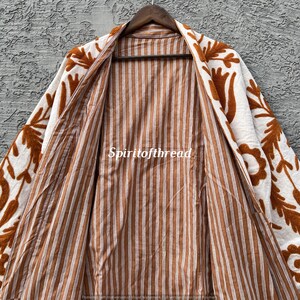 Puede incluir: Chaqueta abierta de color crema con bordados florales marrones en los bordes. El interior presenta un patr&oacute;n de rayas marrones y blancas. El texto "Spiritofthread" es visible en la chaqueta.