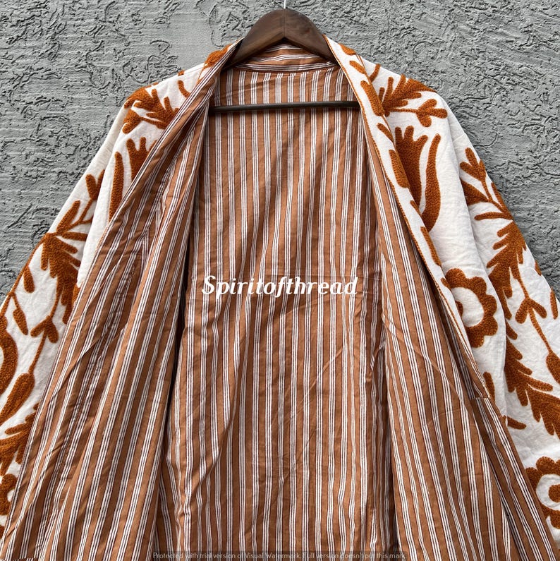 Puede incluir: Chaqueta abierta de color crema con bordados florales marrones en los bordes. El interior presenta un patr&oacute;n de rayas marrones y blancas. El texto "Spiritofthread" es visible en la chaqueta.